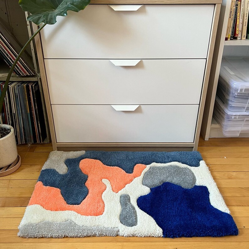 K&ouml;nnte beinhalten: Eine wei&szlig;e Kommode mit drei Schubladen und silbernen Griffen. Die Kommode steht vor einem Teppich mit einem abstrakten Design in Blau, Grau und Orange.