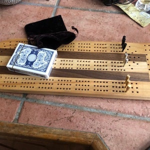 Può includere: Un tabellone da cribbage in legno con intarsi in noce e acero. Il tabellone ha una forma arrotondata ed è fatto di legno chiaro. Ci sono pioli in ottone e un mazzo di carte sul tabellone.