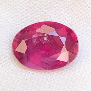Puede incluir: Una gema de rubí de corte ovalado con un tono rojo intenso y una superficie multifacetada. La piedra muestra una claridad interna nítida y facetas geométricas precisas que reflejan la luz, creando un aspecto brillante sobre tela blanca.
