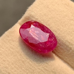 May include: A deep red oval-shaped gemstone with a faceted cut. The gemstone is displayed on a beige velvet surface.