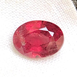 May include: A loose, oval-cut gemstone with a deep, vibrant red color and multiple facets that reflect light. The stone is positioned on a white, textured fabric surface, showcasing its translucent quality and polished, symmetrical shape.