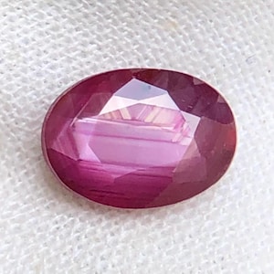May include: An oval-cut gemstone with a deep magenta and purple hue. The stone features a faceted surface that reflects light, creating a clear pattern of geometric shapes across its polished, translucent, and vibrant colored surface.