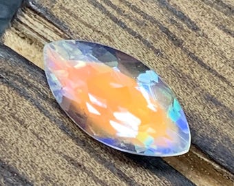 May include: A marquise-cut gemstone with a vibrant, iridescent display of colors, including orange, blue, and hints of green. The gemstone is set against a backdrop of a dark, textured wooden surface.