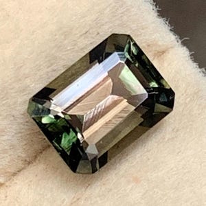 以下が含まれることがあります： 緑と茶色の色合いが混ざった長方形のエメラルドカットの宝石。石のファセット加工された表面は光を反射し、きらめく効果を生み出します。宝石は柔らかいオフホワイトの背景に置かれ、その色と透明度を際立たせています。