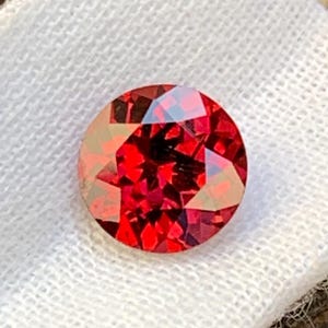 Puede incluir: Una gema roja, redonda y facetada, descansa sobre una tela blanca. La gema tiene un corte brillante, que refleja la luz para crear un efecto brillante. La gema se encuentra sobre un fondo de superficie de madera.