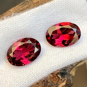 May include: Two oval-cut, faceted gemstones with a deep red hue. The gems are displayed on a white cloth, showcasing their brilliance. The stones are likely garnets, perfect for jewelry making.