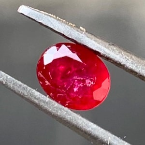 Pode incluir: Uma gema oval de cor vermelha vibrante segurada por uma pinça. A gema é lisa e polida, refletindo a luz.