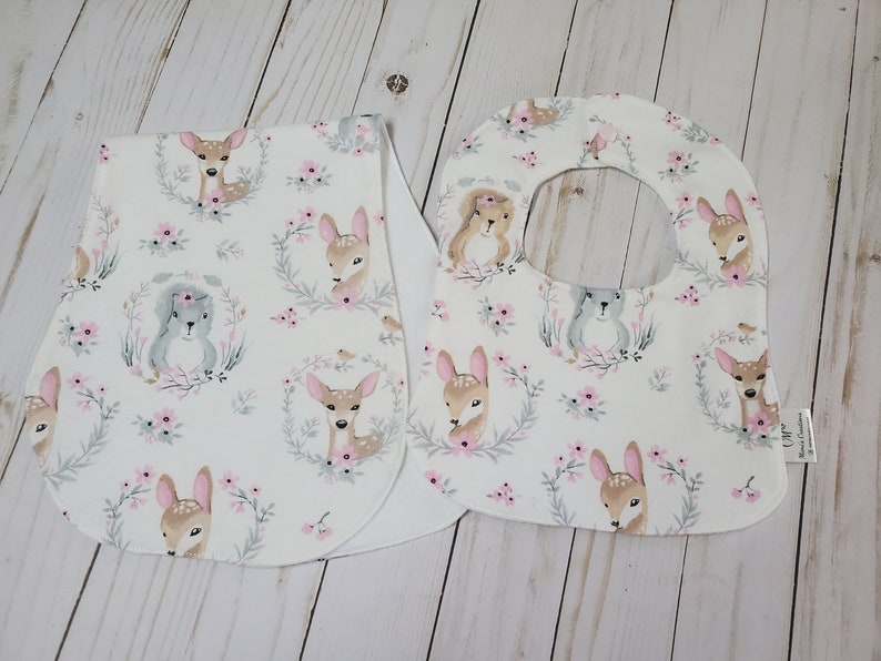 May include: Two white baby bibs with a woodland animal print. The print features deer, squirrels, and pink flowers on a white background.