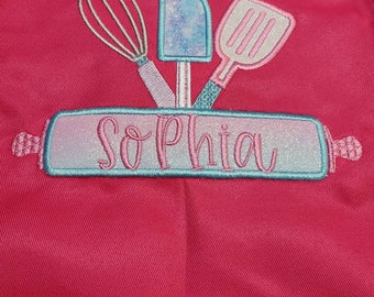 May include: Pink child's apron with a blue and pink embroidered design of kitchen utensils and the name "Sophia" on a rolling pin.