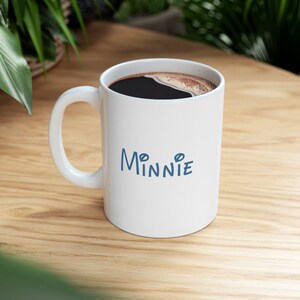 Puede incluir: Taza de caf&eacute; de cer&aacute;mica blanca con caf&eacute; negro en su interior y la palabra "Minnie" escrita en azul en la parte delantera.
