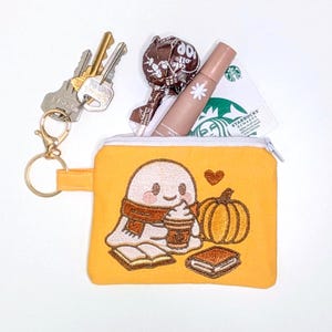 May include: A yellow zipped pouch featuring an embroidered ghost design, a pumpkin, and books. The pouch contains keys, a lollipop, lip gloss, and a Starbucks rewards card. The ghost is wearing a scarf and holding a coffee cup.