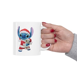 Puede incluir: Taza de cer&aacute;mica blanca con un personaje de dibujos animados azul y rojo que lleva un gorro de Pap&aacute; Noel y sostiene un juguete. El personaje es Stitch de la pel&iacute;cula de Disney Lilo & Stitch.