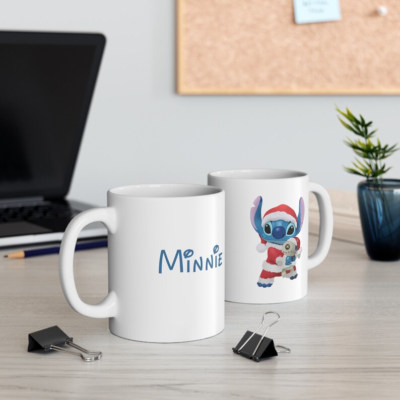 Puede incluir: Taza de cer&aacute;mica blanca con un personaje azul Stitch que lleva un gorro de Pap&aacute; Noel y sostiene un hombre de jengibre. La taza tiene la palabra "Minnie" escrita en azul.