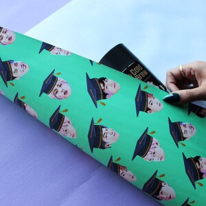 May include: A roll of green wrapping paper with a repeating pattern of a person wearing a graduation cap. The person's face is visible and the cap is black with a gold tassel.