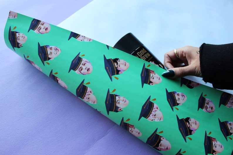 May include: A roll of green wrapping paper with a repeating pattern of a person wearing a graduation cap. The person's face is visible and the cap is black with a gold tassel.