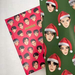 May include: A red notebook with a repeating pattern of a man's face wearing a black hat. The notebook has the word "maniac" printed in green letters. The notebook is lying on top of green wrapping paper with a repeating pattern of a man's face wearing a red Santa hat.