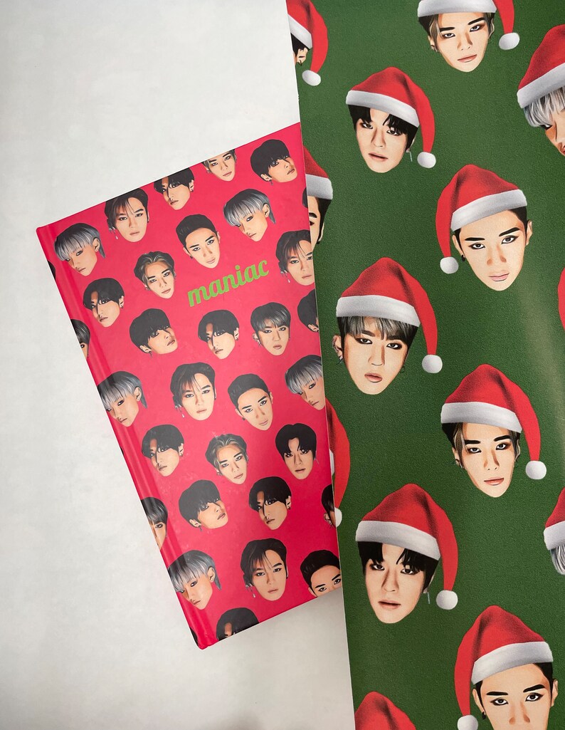 May include: A red notebook with a repeating pattern of a man's face wearing a black hat. The notebook has the word "maniac" printed in green letters. The notebook is lying on top of green wrapping paper with a repeating pattern of a man's face wearing a red Santa hat.