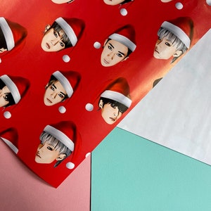 May include: Red wrapping paper with a repeating pattern of a person's face wearing a Santa hat. The person has black hair and is looking directly at the viewer.
