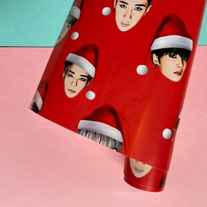 May include: Red wrapping paper with a repeating pattern of men's faces wearing Santa hats. The faces are all different, but they all have the same expression.