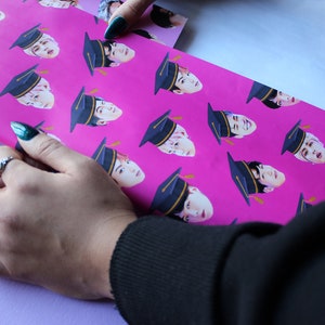 May include: Pink wrapping paper with a repeating pattern of a person's face wearing a graduation cap. The paper is being folded by a hand wearing a ring.