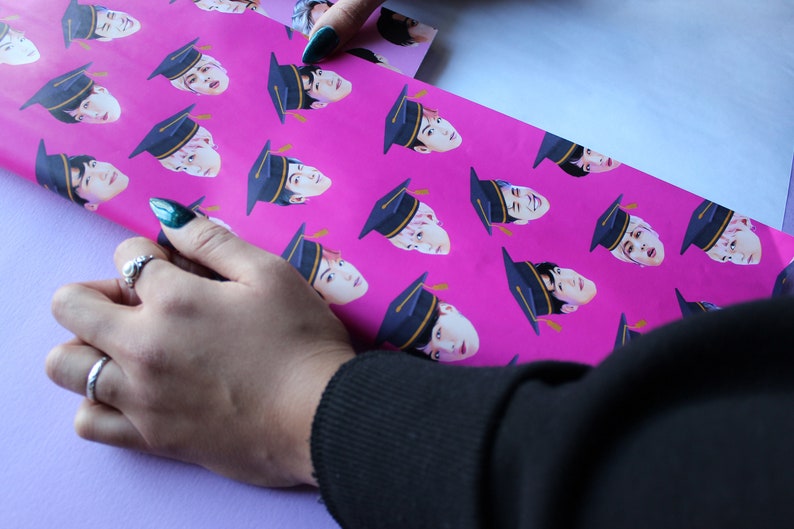 May include: Pink wrapping paper with a repeating pattern of a person's face wearing a graduation cap. The paper is being folded by a hand wearing a ring.
