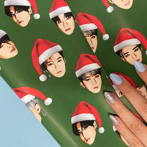 May include: Green wrapping paper with a repeating pattern of cartoon faces wearing Santa hats. The faces are all different, but they all have the same expression.