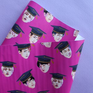 May include: Pink wrapping paper with a repeating pattern of graduation caps and faces of young men. The caps are black with gold trim. The faces are all different, but they are all smiling.