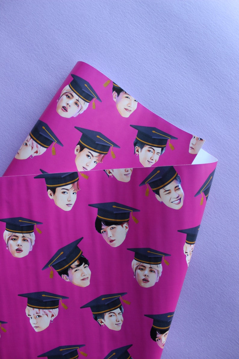 May include: Pink wrapping paper with a repeating pattern of graduation caps and faces of young men. The caps are black with gold trim. The faces are all different, but they are all smiling.