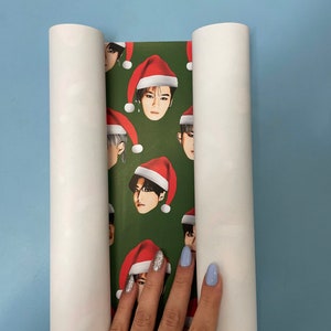 May include: A roll of green wrapping paper with a repeating pattern of a man's face wearing a Santa hat. The paper is partially rolled up and a hand is holding the paper.