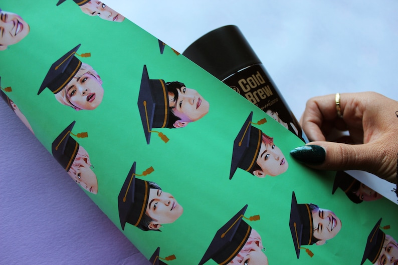 May include: Green wrapping paper with a repeating pattern of a person's face wearing a black graduation cap. The paper is being held by a hand with green nail polish.