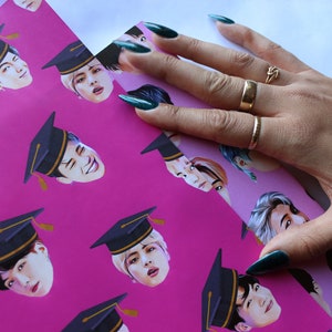 May include: Pink wrapping paper with a repeating pattern of a man's face wearing a graduation cap. The man has black hair and is wearing a black and gold graduation cap. The paper is being held by a hand with green nail polish.
