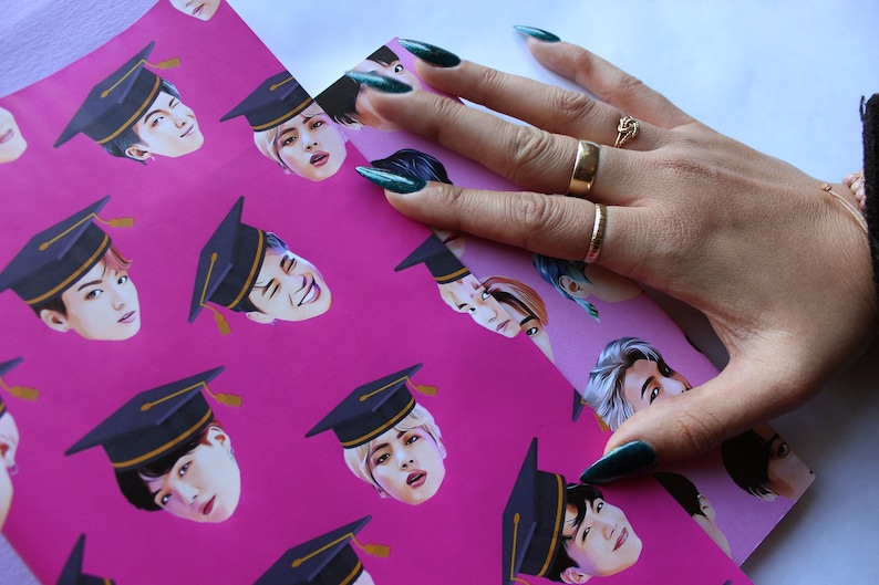 May include: Pink wrapping paper with a repeating pattern of a man's face wearing a graduation cap. The man has black hair and is wearing a black and gold graduation cap. The paper is being held by a hand with green nail polish.