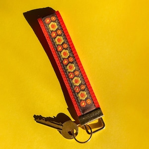 May include: A red and brown key wristlet with a floral pattern in yellow and orange. The wristlet is attached to a key, all set against a bright yellow background. The wristlet is made of fabric and has a metal clasp.
