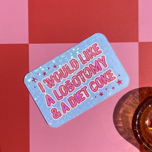 May include: A rectangular sticker with the text "I WOULD LIKE A LOBOTOMY & A DIET COKE" in pink and white. The sticker has a light blue background with a glittery effect and small pink stars. The sticker is on a pink and red surface.