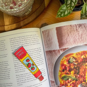 May include: A red and yellow enamel pin shaped like a tube of tomato paste. The pin has the words "Tomato Paste" and "Fine Italian Ingredients" on it. The pin is 4.5 inches long.
