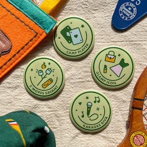 May include: Four green and white round buttons with different designs and text. The buttons feature a campfire with marshmallows, a karaoke microphone, a deck of cards, and a purse and dress. The text on the buttons reads "S'mores Savant", "Karaoke Diva", "Camp Player", and "Glamp Icon".