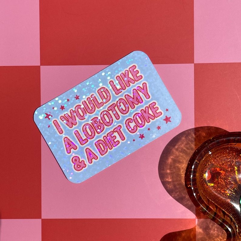 May include: A rectangular sticker with the text "I WOULD LIKE A LOBOTOMY & A DIET COKE" in pink and white. The sticker has a light blue background with a glittery effect and small pink stars. The sticker is on a pink and red surface.