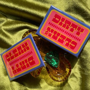 May include: Two rectangular, blue and pink stickers with the words "DIRTY" and "CLEAN" in orange, are displayed. The stickers are on a clear, amber-colored glass dish, with a green heart-shaped object. The background is a green, satin fabric.