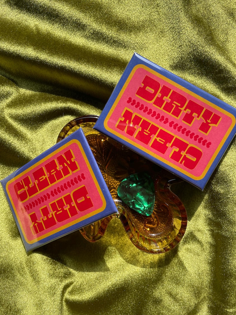May include: Two rectangular, blue and pink stickers with the words "DIRTY" and "CLEAN" in orange, are displayed. The stickers are on a clear, amber-colored glass dish, with a green heart-shaped object. The background is a green, satin fabric.