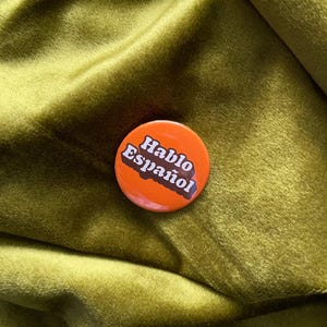 May include: A round, orange button with the words "Hablo Español" in white and brown text. The button is set against a textured, olive-green background.