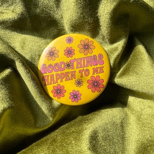 May include: A circular yellow pin-back button featuring the pink text GOOD THINGS HAPPEN TO ME in a stylized font. The design is decorated with multiple small orange and pink flowers scattered across the surface of the round button.