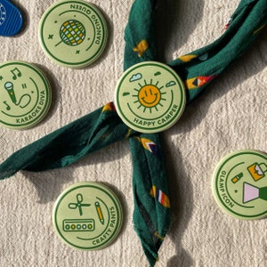 May include: Five green and yellow round buttons with different designs and text. The buttons feature a disco ball, a microphone, a smiling sun, a pair of scissors, a pencil, and a paint palette. The text on the buttons reads "Dancing Queen", "Karaoke Diva", "Happy Camper", "Crafty Pants", and "Glamp Icon".