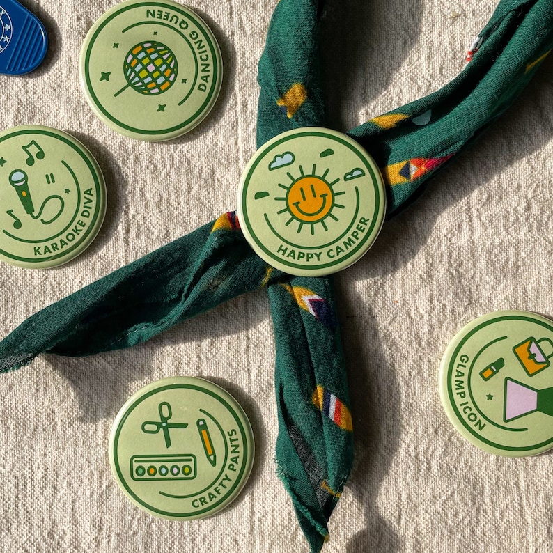 May include: Five green and yellow round buttons with different designs and text. The buttons feature a disco ball, a microphone, a smiling sun, a pair of scissors, a pencil, and a paint palette. The text on the buttons reads "Dancing Queen", "Karaoke Diva", "Happy Camper", "Crafty Pants", and "Glamp Icon".
