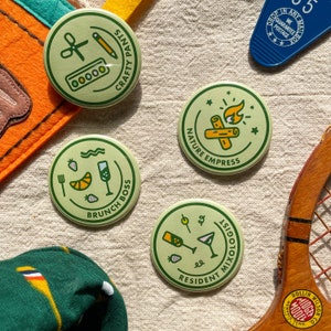 May include: Four green and yellow round buttons with different designs. The buttons feature illustrations of a paint palette, a campfire, a cocktail, and a brunch plate. The text on the buttons reads "Crafty Pants", "Nature Empress", "Resident Mixologist", and "Brunch Boss".