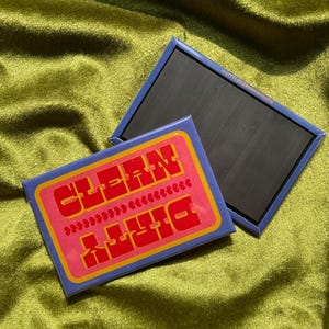 May include: Two rectangular magnets with a retro design. One magnet features the words "CLEAN" and "DIRTY" in red, with a pink background and yellow border. The other magnet is plain black with a blue border. The background is a textured, olive green fabric.