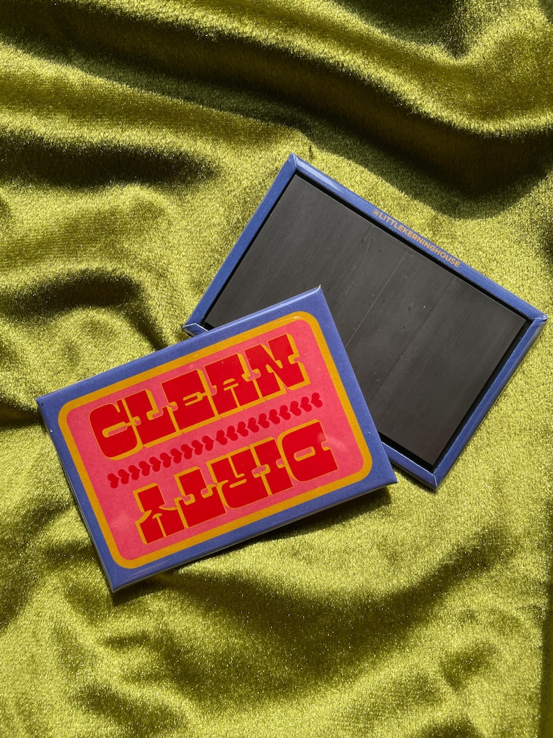 May include: Two rectangular magnets with a retro design. One magnet features the words "CLEAN" and "DIRTY" in red, with a pink background and yellow border. The other magnet is plain black with a blue border. The background is a textured, olive green fabric.