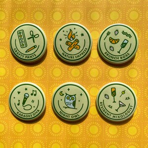 May include: Six round, green and cream colored buttons with different illustrations and text. The buttons feature illustrations of a campfire, a microphone, a cocktail, a pair of scissors, an owl, and a lemon. The text on the buttons reads "Crafty Pants", "Nature Empress", "Brunch Boss", "Karaoke Diva", "Night Owl", and "Resident Mixologist".