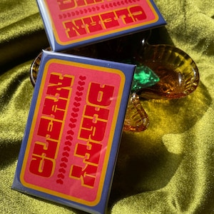 May include: Two rectangular magnets with the words "DIRTY" and "CLEAN" in red block letters on a pink background. The magnets have a blue border and a yellow outline. One magnet is slightly behind the other. The background is a green, textured fabric.