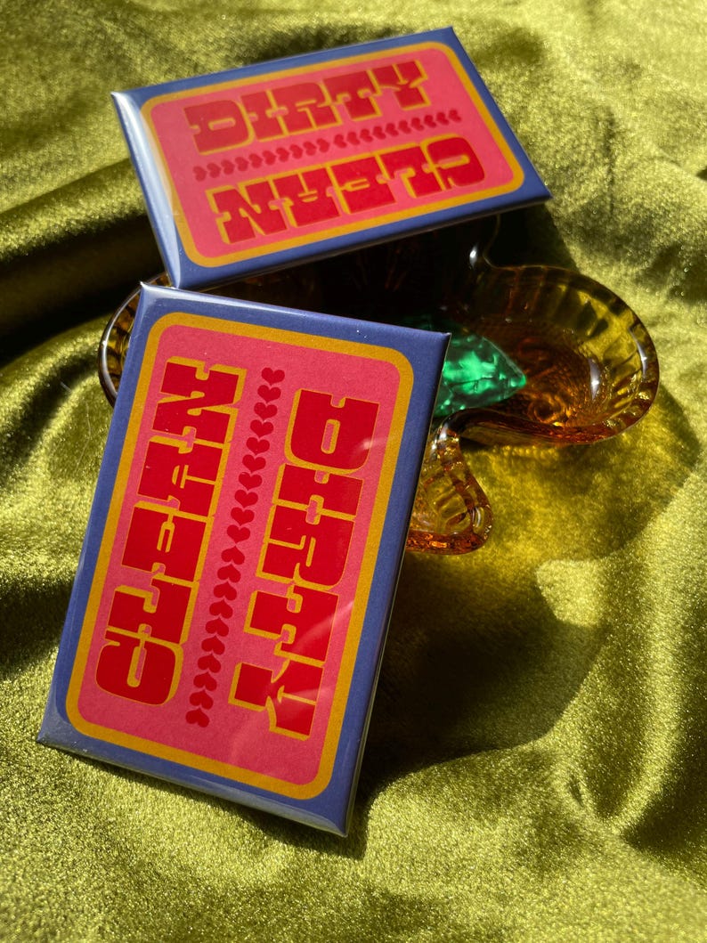 May include: Two rectangular magnets with the words "DIRTY" and "CLEAN" in red block letters on a pink background. The magnets have a blue border and a yellow outline. One magnet is slightly behind the other. The background is a green, textured fabric.