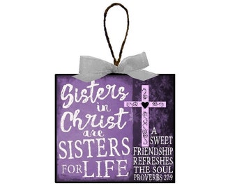 May include: A purple and black wooden sign with a white cross and the text "Sisters in Christ are Sisters for Life" and a quote from Proverbs 27:9, "A sweet friendship refreshes the soul."